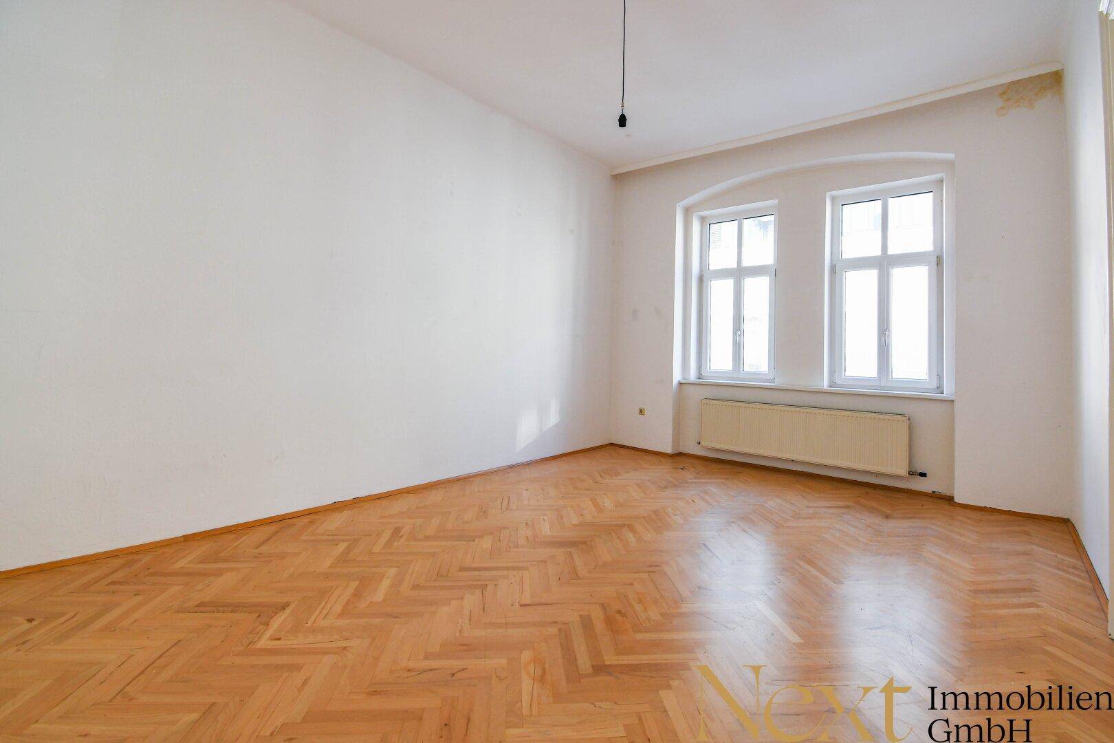 Geräumiges Zimmer mit Fischgrätparkett und zwei großen Fenstern, ideal als Schlaf- oder Wohnbereich.