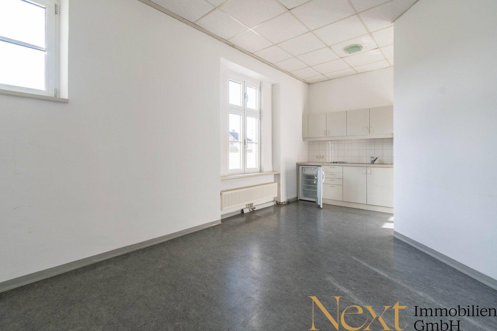 Kleine, funktionale Küchenzeile mit weißen Schränken, Spüle und Kühlschrank in einem hellen Raum mit Fenster.