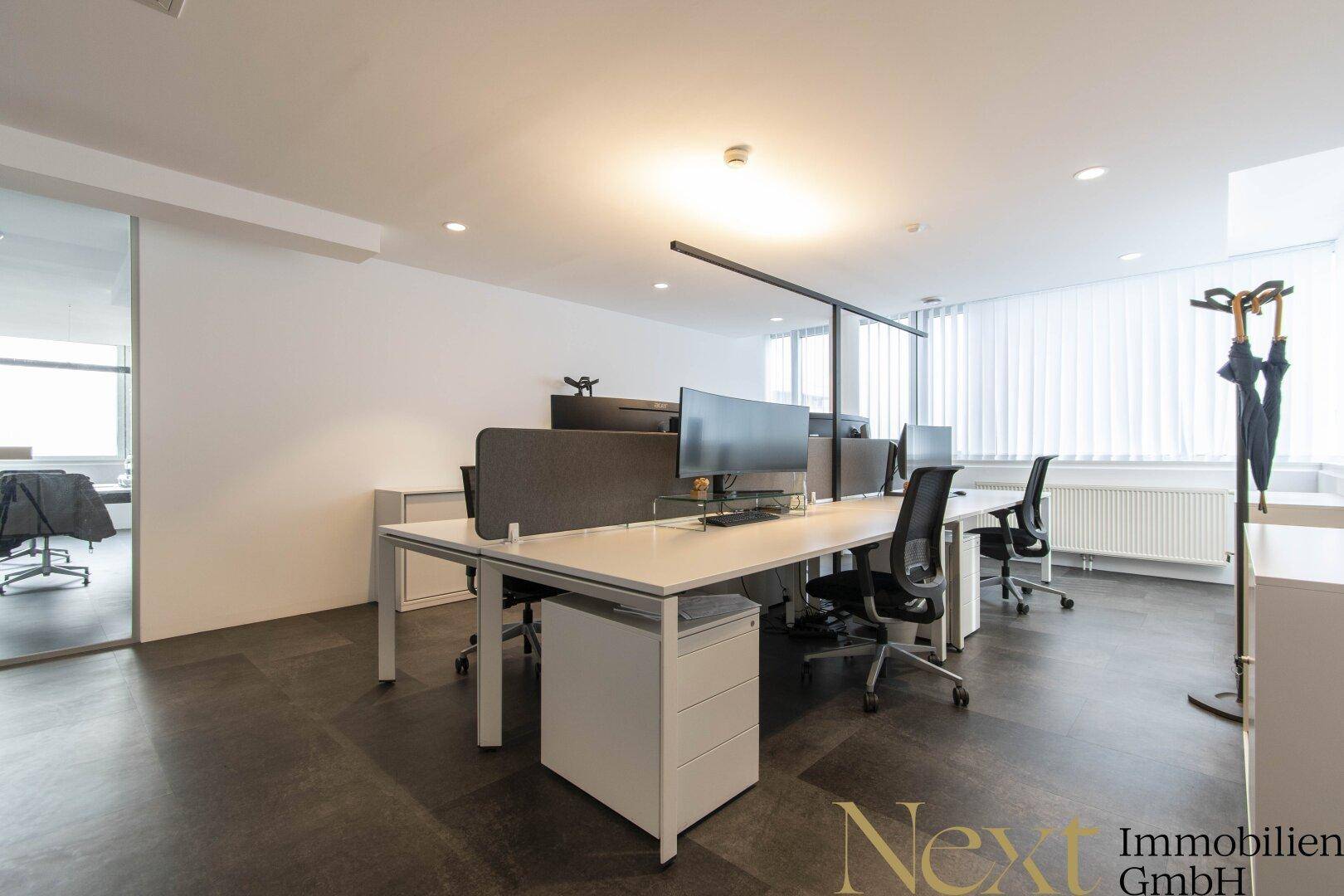 Moderner Bürobereich mit mehreren Schreibtischen, ergonomischen Stühlen und großen Fenstern mit Jalousien.