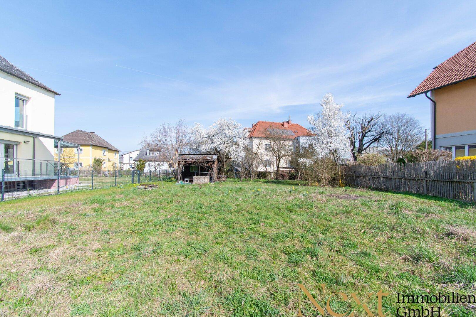 Sonniges Gartengrundstück mit viel Grünfläche, einem Holzschuppen und umgebenden Häusern.
