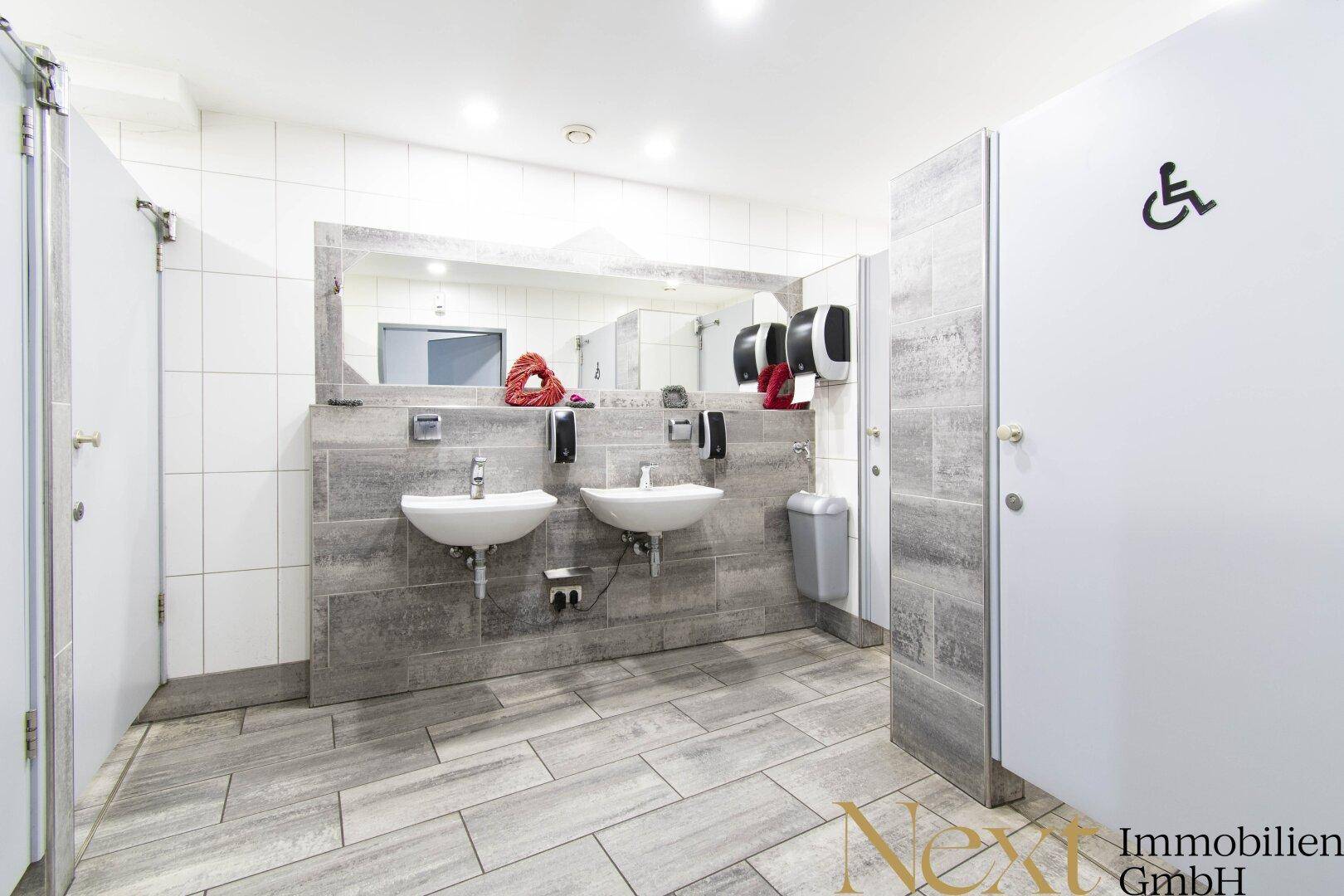 Moderner öffentlicher Waschraum mit zwei Waschbecken, großen Spiegeln und barrierefreier Toilette.
