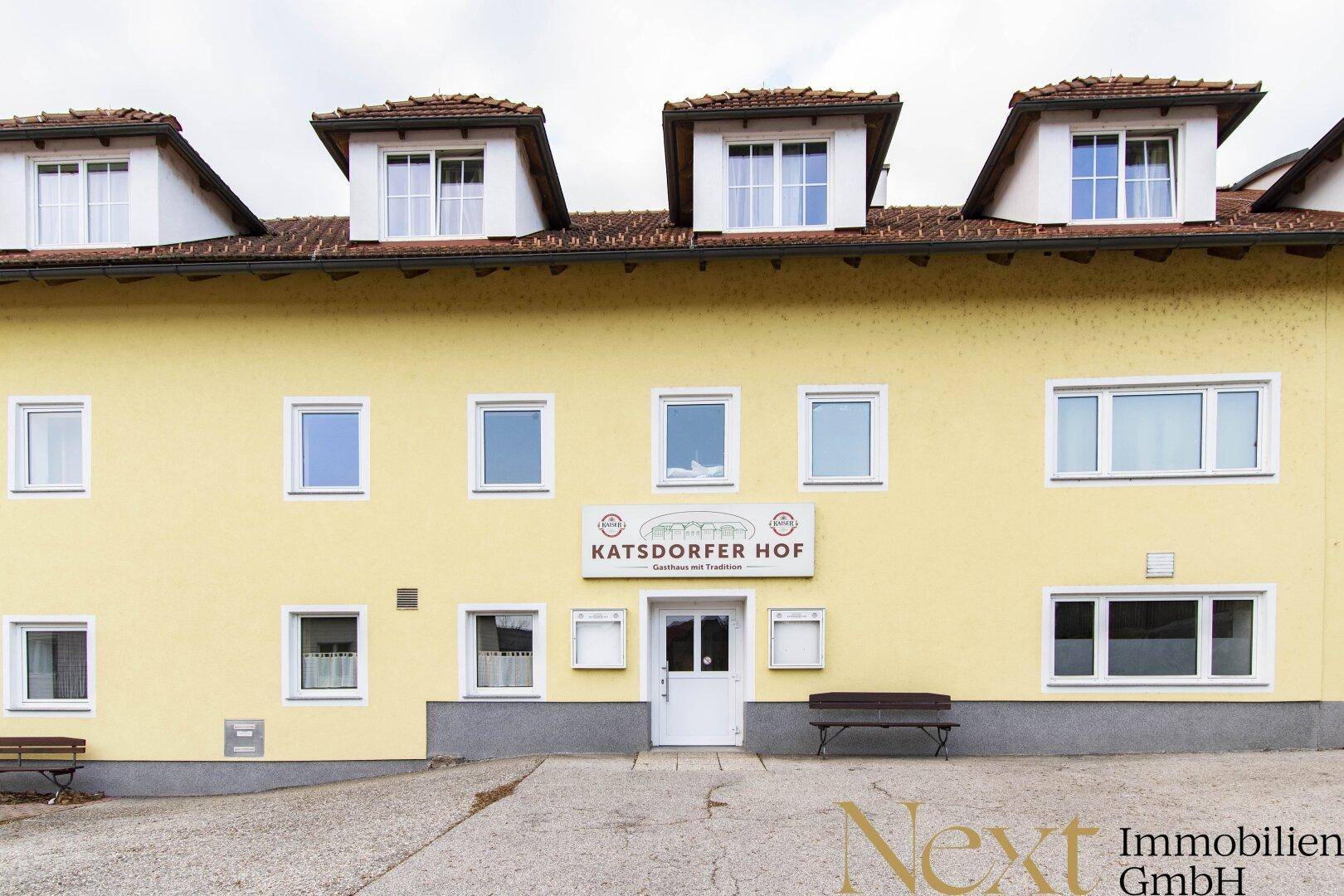 Traditionelles Gebäude mit gelber Fassade, weißen Fenstern und mehreren Dachgauben, inklusive Eingangsbereich mit Sitzbänken.