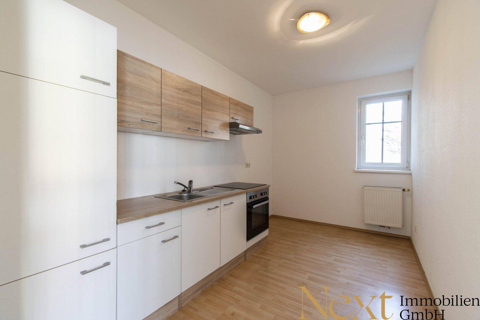 Helle, unmöblierte Küche mit weißen und holzfarbenen Schränken, Herd und Fenster.