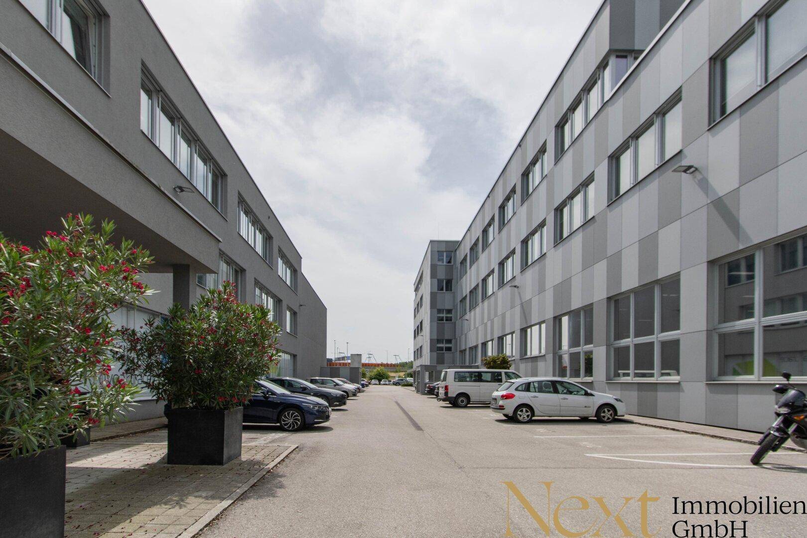 Moderne Geschäftsgebäude mit grauen Fassaden und großen Fenstern, umgeben von einem Parkplatz mit Fahrzeugen und dekorativen Pflanzen.