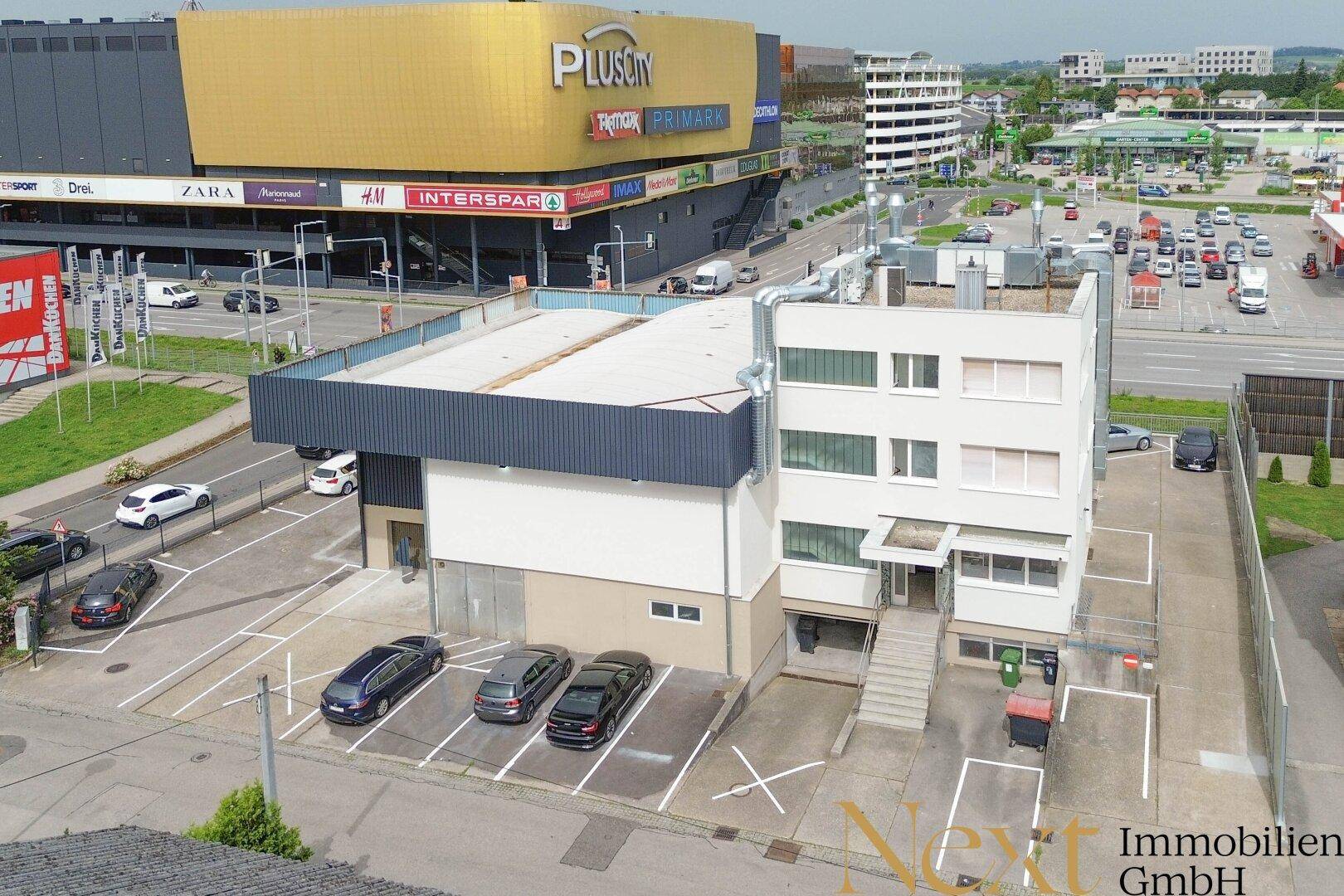 Luftaufnahme eines Geschäftsgebäudes mit Parkplätzen und einem Einkaufszentrum im Hintergrund.