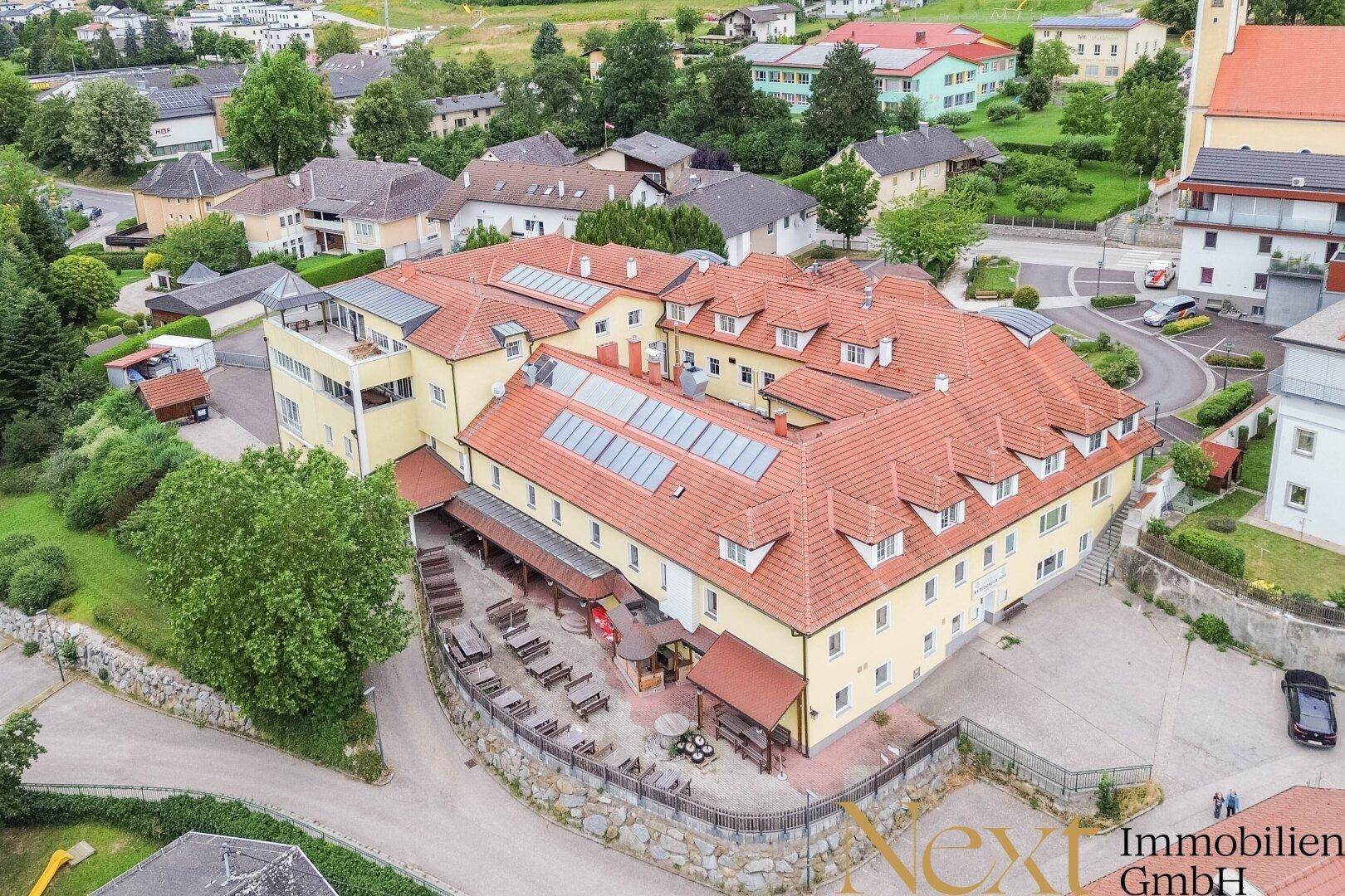 Luftaufnahme eines großen Gebäudekomplexes mit roten Dächern, Solaranlagen und Außengastronomiebereich.
