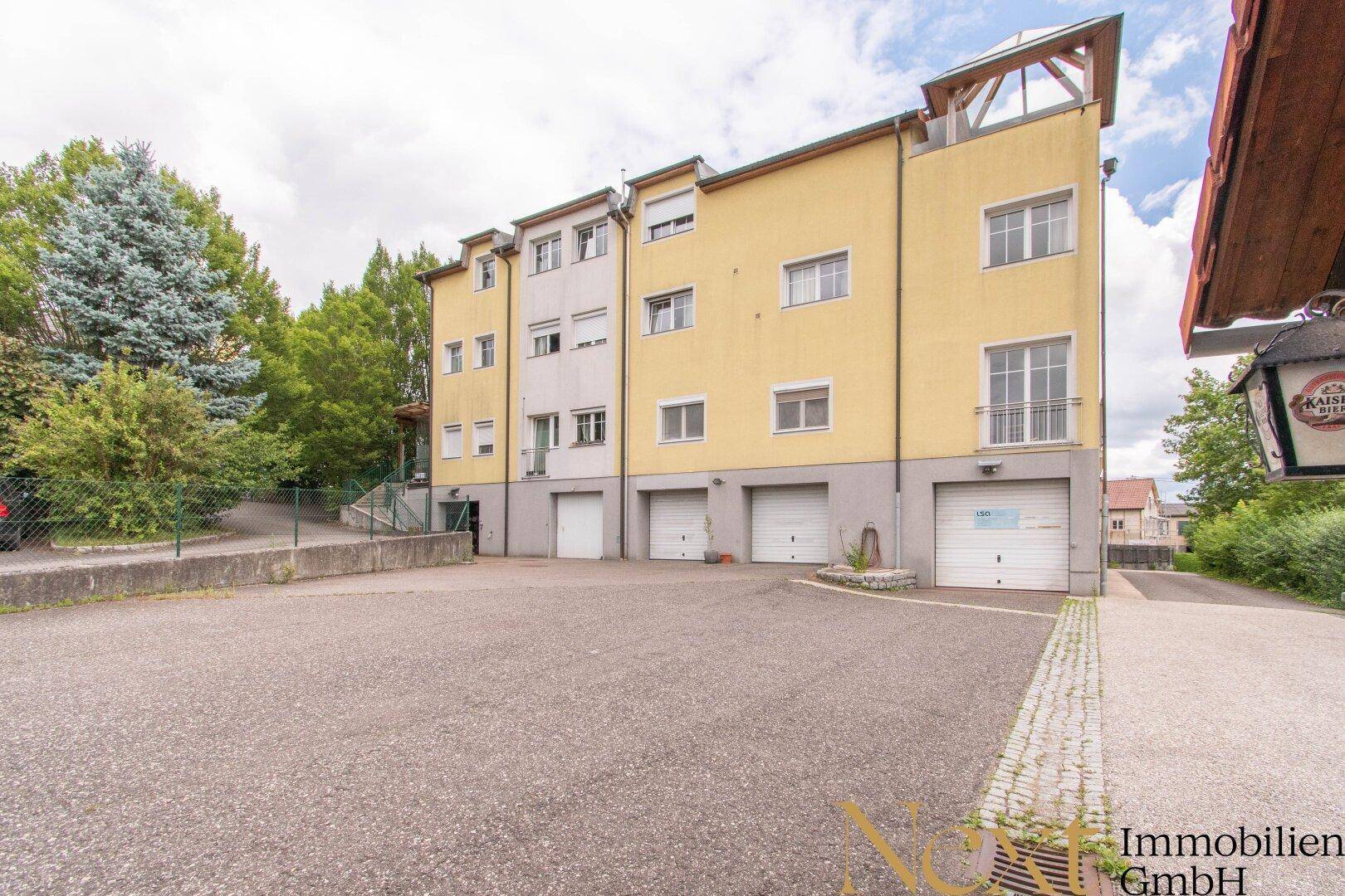 Gelbe Gebäudefassade mit Garagen im Erdgeschoss und einem großen gepflasterten Parkplatz davor.
