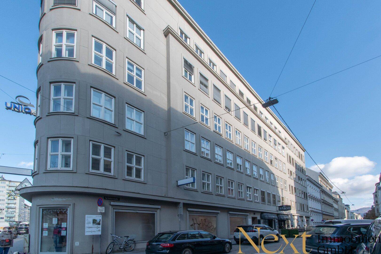 Graues Eckgebäude mit vielen Fenstern und Geschäften im Erdgeschoss an einer städtischen Straße.