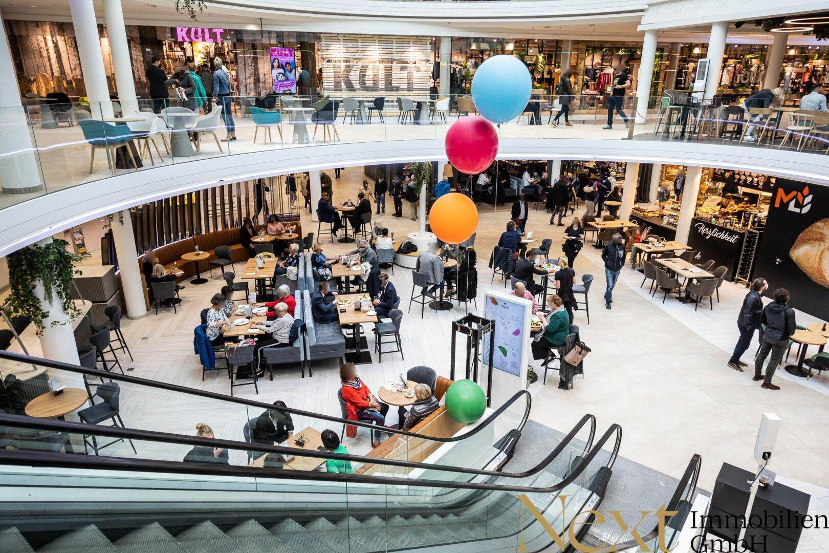 Belebtes Einkaufszentrum mit einem Food-Court auf mehreren Ebenen, vielen Besuchern und Rolltreppen.