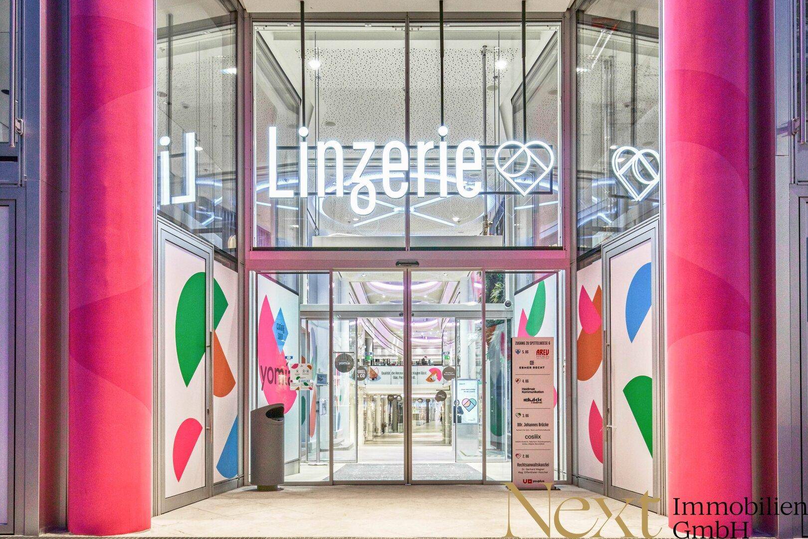 Moderner Eingangsbereich eines Geschäfts mit großen Glastüren, leuchtenden Neonschildern und farbenfrohen Grafiken.