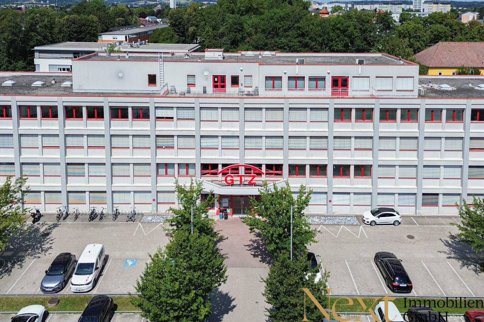 Luftaufnahme eines großen Bürogebäudes mit vielen Fenstern, einem Parkplatz und Bäumen im Vordergrund.