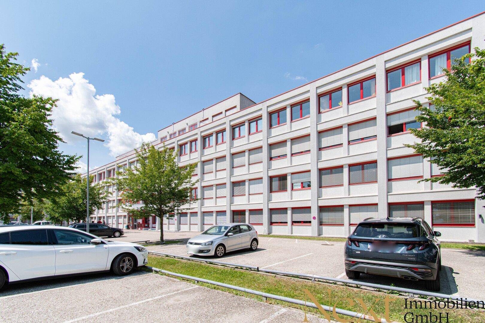 Moderne Bürogebäude-Fassade mit vielen Fenstern und davor liegenden Parkplätzen unter blauem Himmel.