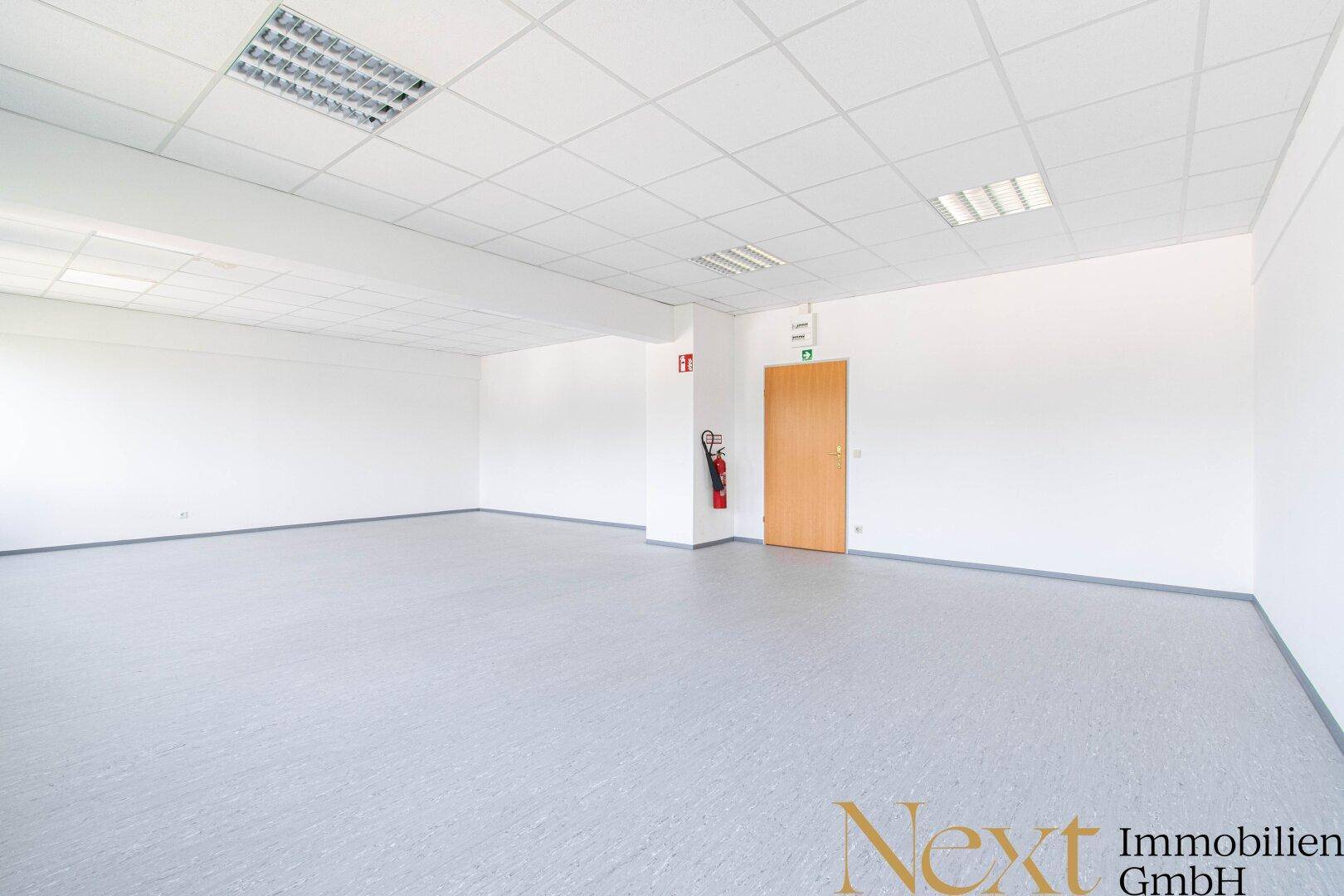 Weitläufiger, leerer Bürobereich mit hellem Vinylboden und einer Holztür am Ende des Raumes.
