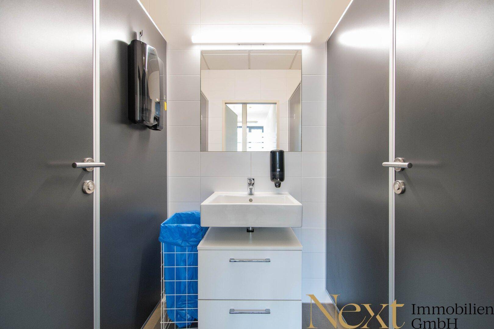 Saubere Toilettenanlage mit Waschbecken, Spiegel und zwei Kabinentüren, in modernem Design.
