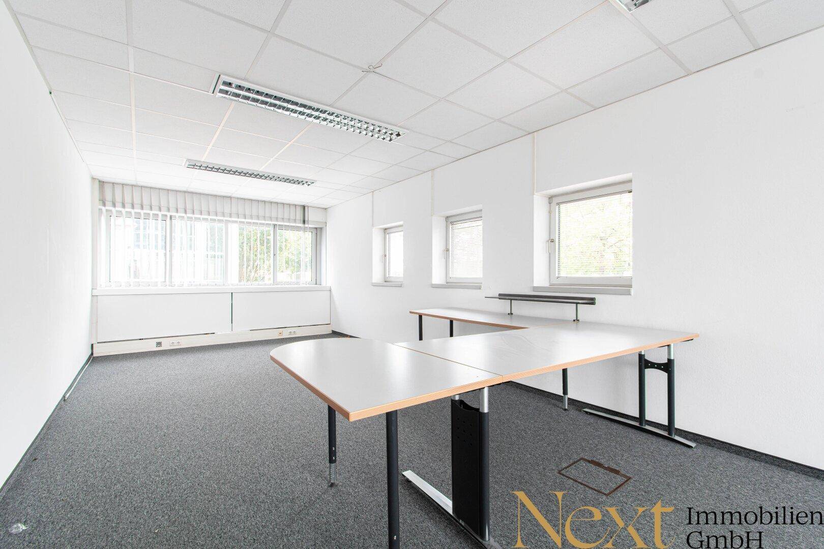 Helles Büro mit grauem Teppichboden, weißen Wänden und zwei Schreibtischen am Fenster.