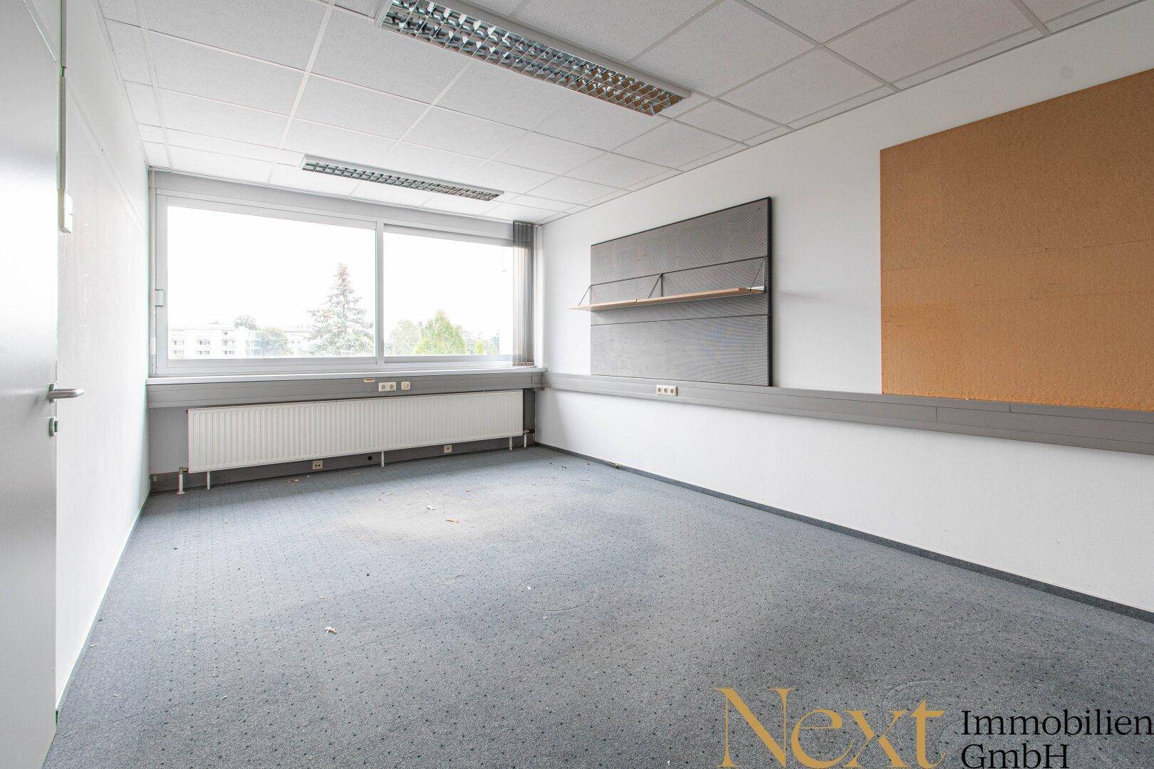 Leeres Büro mit grauem Teppichboden, großem Fenster und Heizkörper unter der Fensterbank.