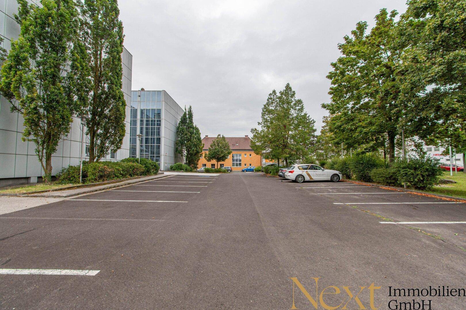 Außenparkplatz mit Bäumen und weiteren Gebäuden im Hintergrund, bietet ausreichend Stellflächen.