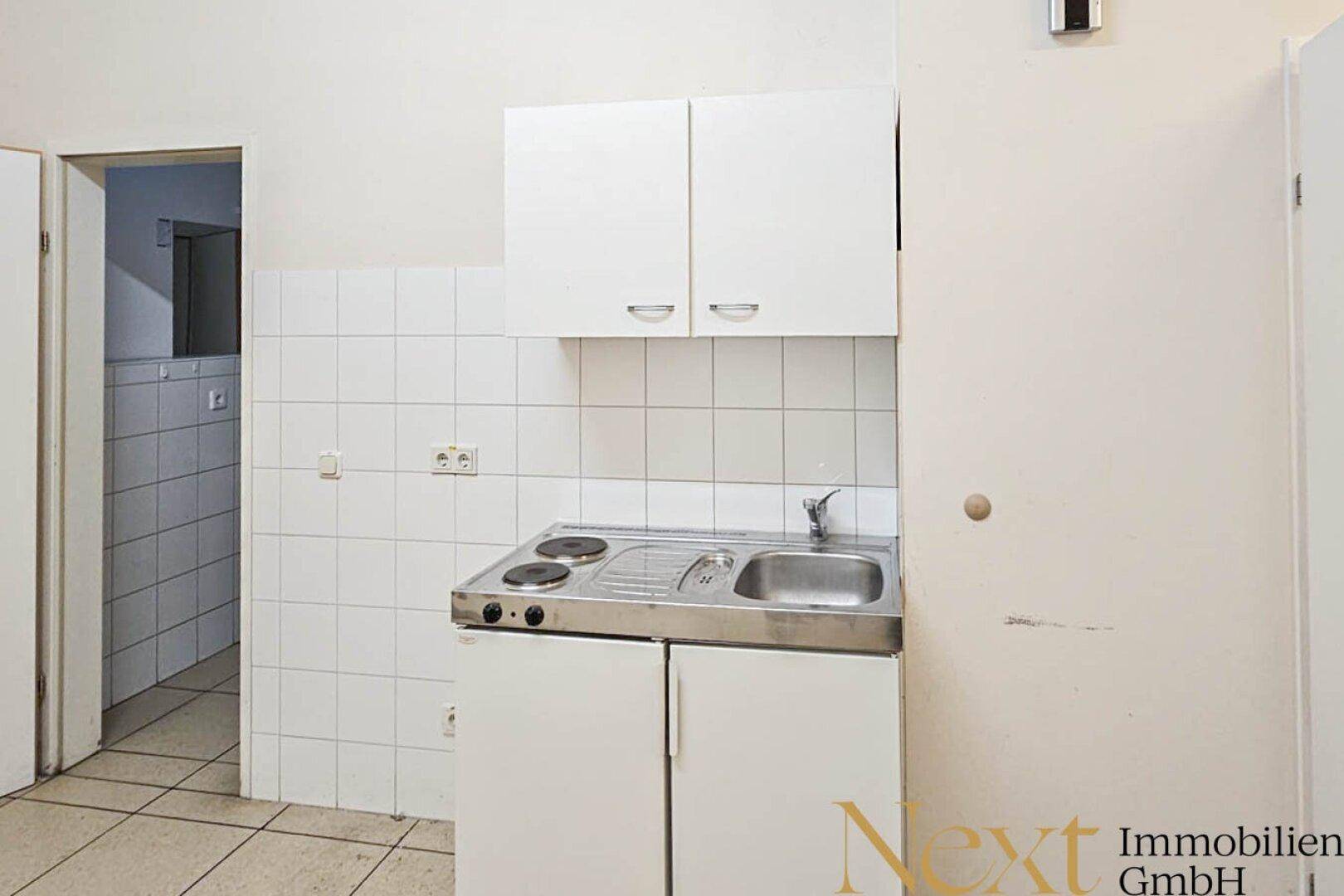 Kleine Küchenzeile mit Spüle, zwei Kochplatten und weißen Hängeschränken, ideal für Personalräume.