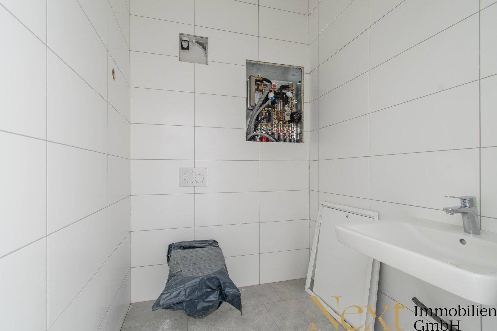 Gefliester Sanitärraum mit Waschbecken und vorbereiteter Toiletteninstallation, noch im Ausbau.