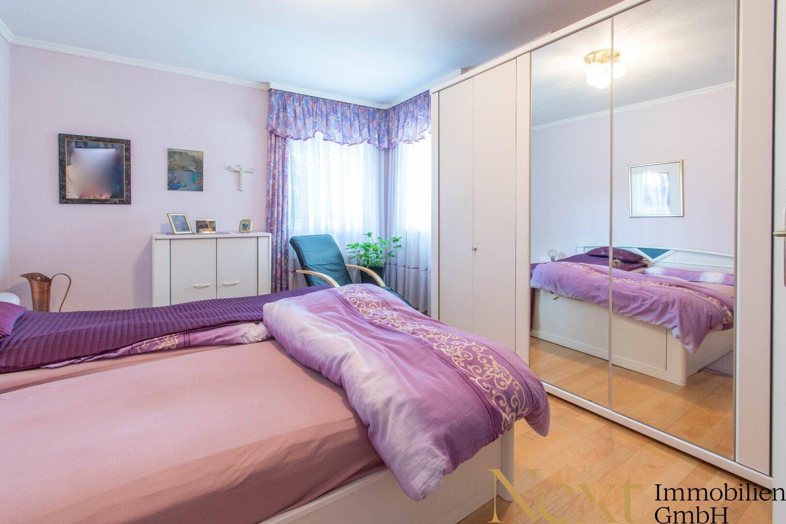 Schlafzimmer mit Doppelbett, Spiegelschrank und lila Bettwäsche.