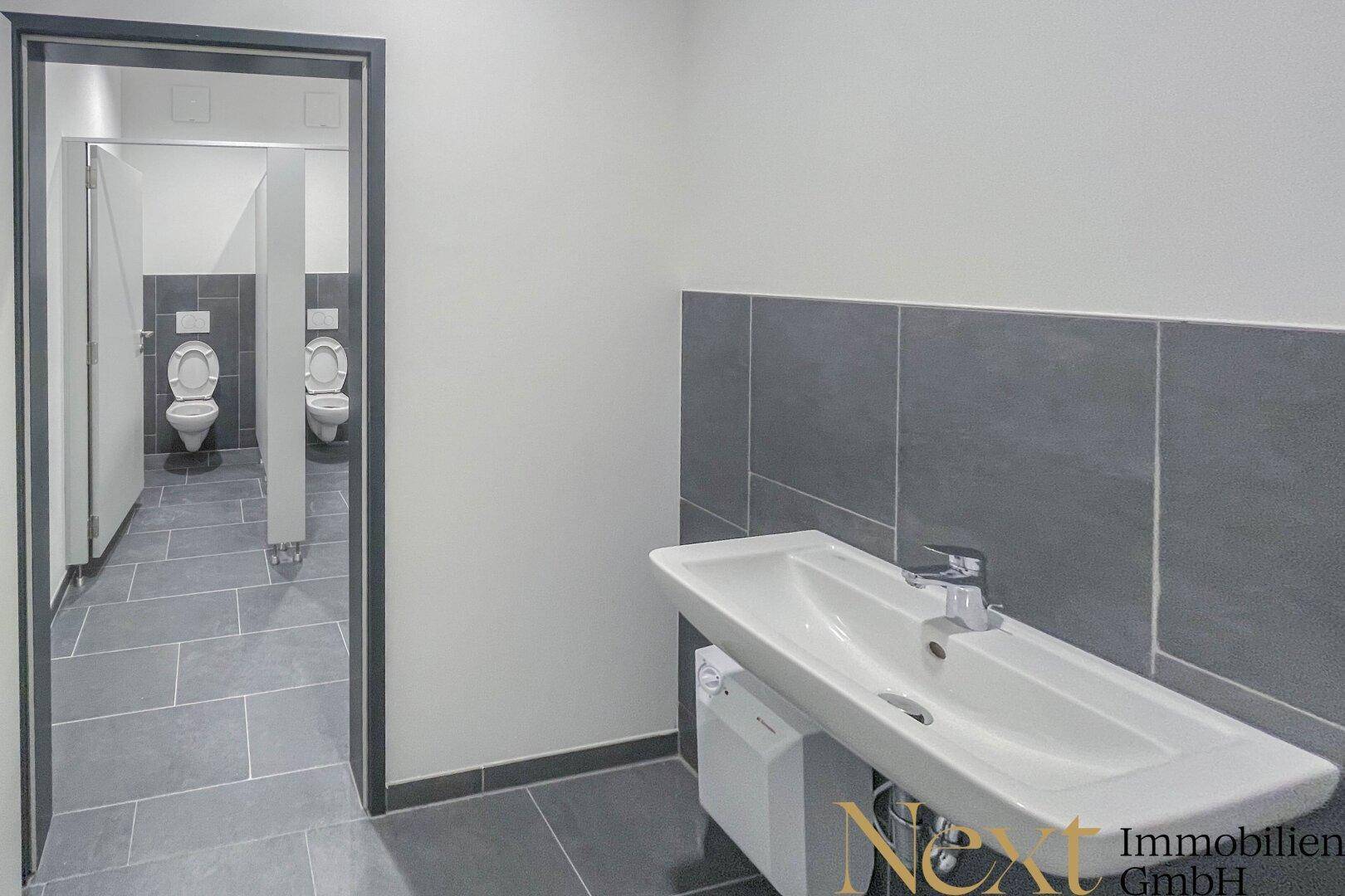 Modernes Badezimmer mit Waschbecken und mehreren Toilettenkabinen, ausgestattet mit grauen Fliesen.