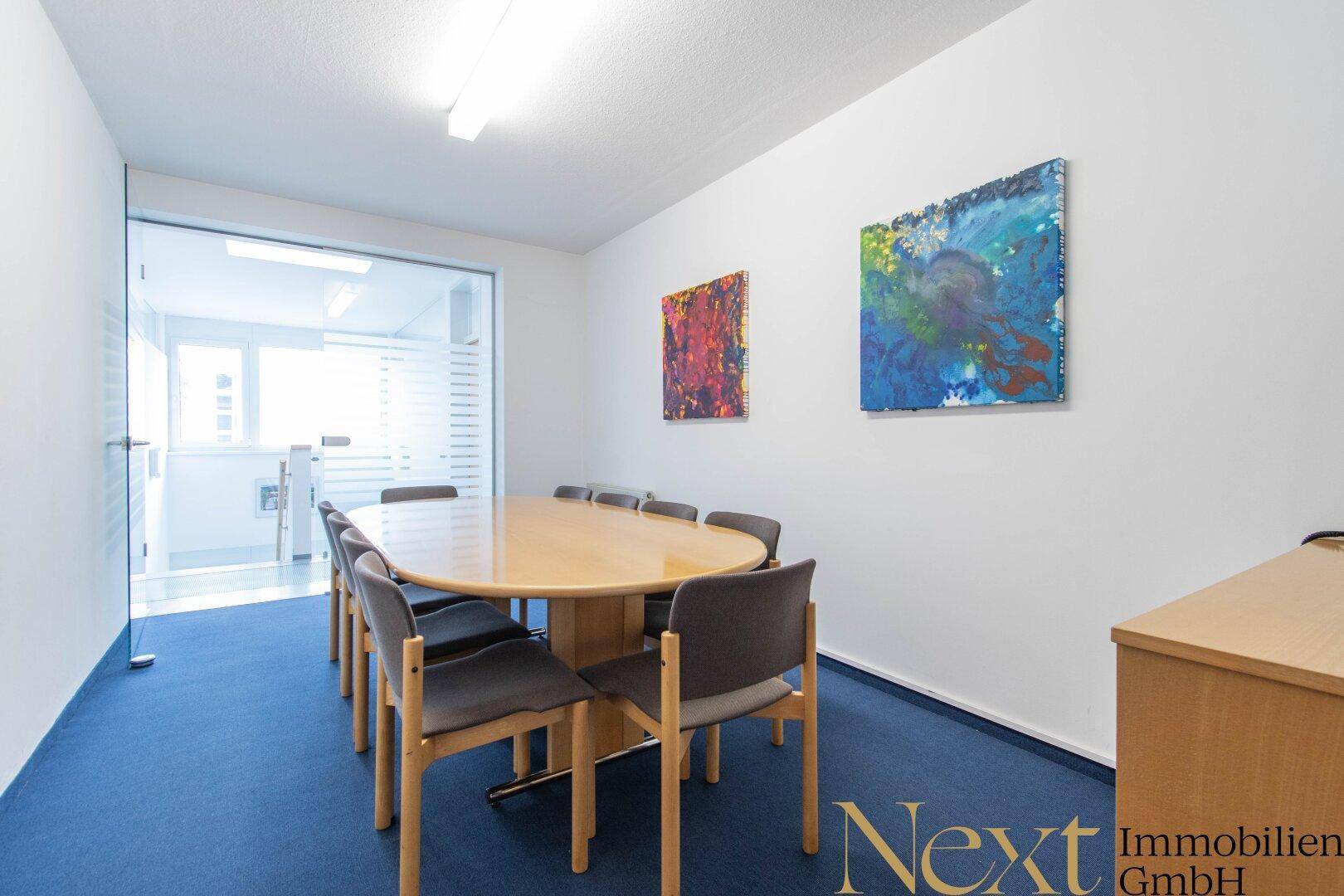 Konferenzraum mit großem ovalen Tisch und Stühlen, durch eine Glastür von einem weiteren Raum getrennt.