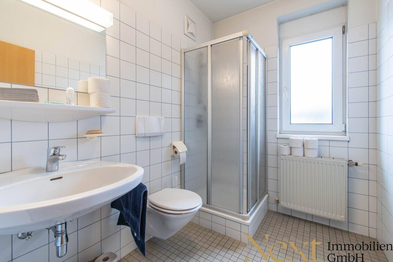 Helles Badezimmer mit Duschkabine, WC und Waschbecken, komplett gefliest und mit Fenster für Tageslicht.