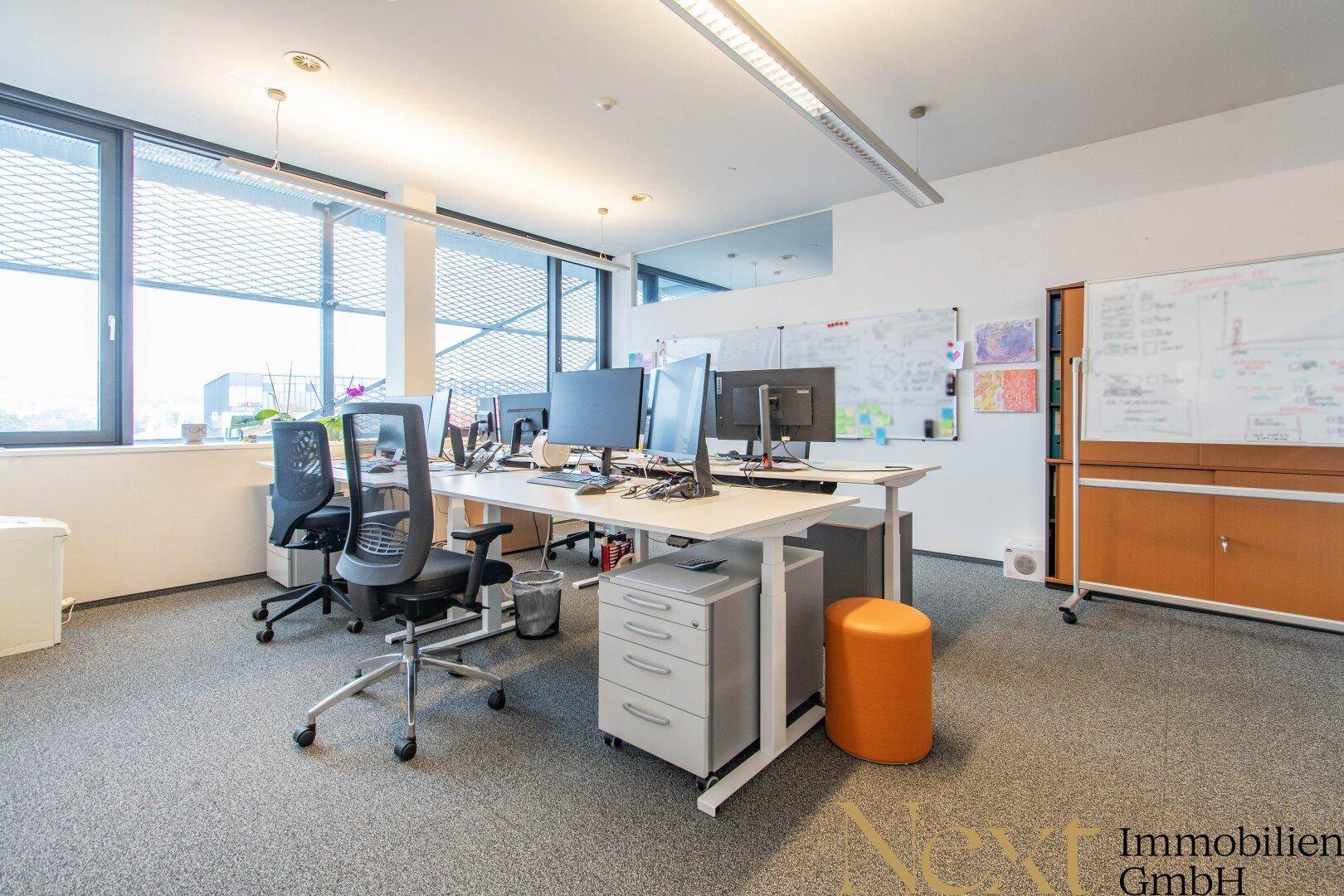 Großraumbüro mit mehreren Arbeitsplätzen, ergonomischen Stühlen und viel Tageslicht durch große Fenster.