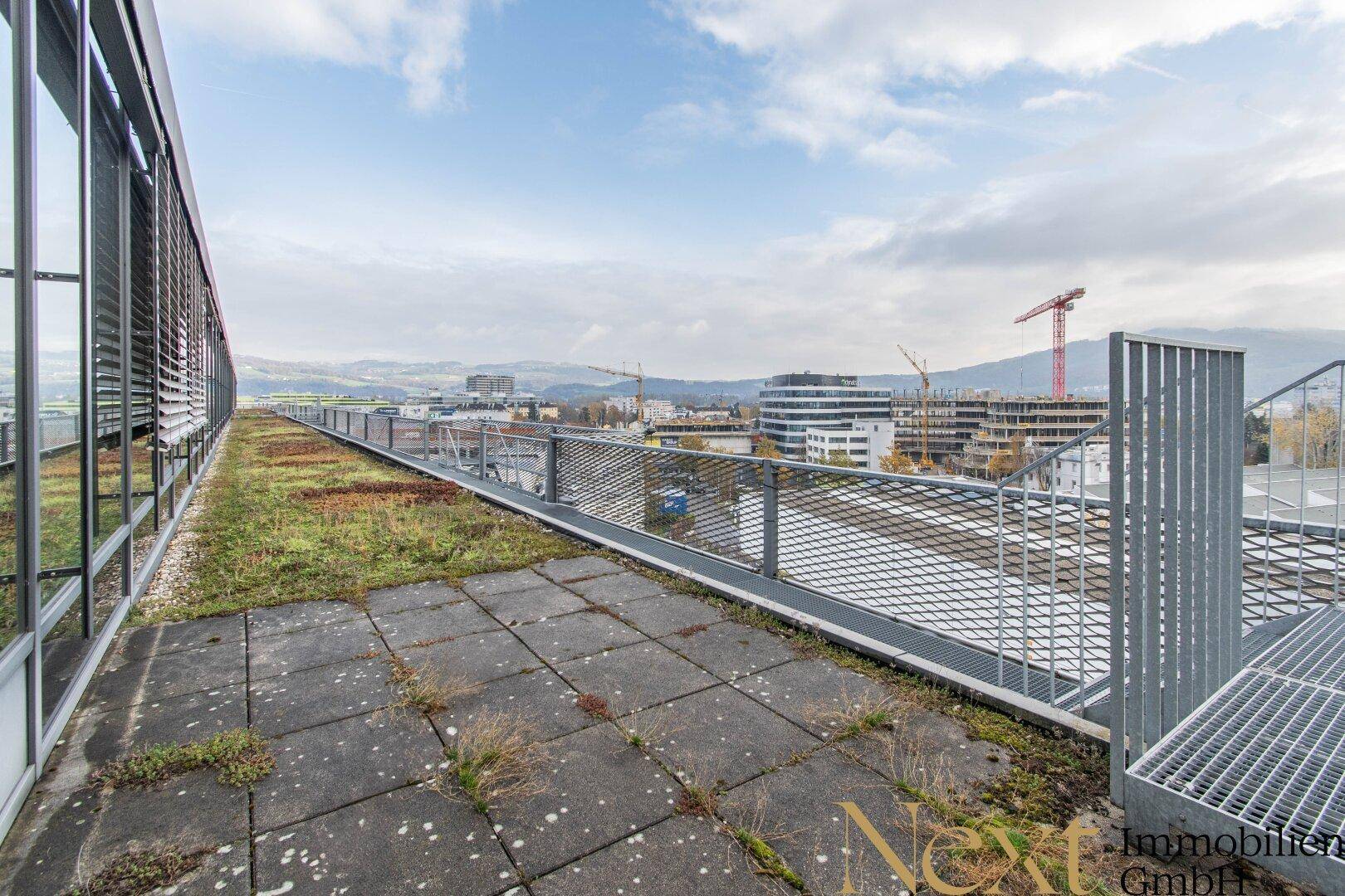 Lange Dachterrasse mit begrüntem Bereich und Blick auf die umliegende Stadtlandschaft und Berge.