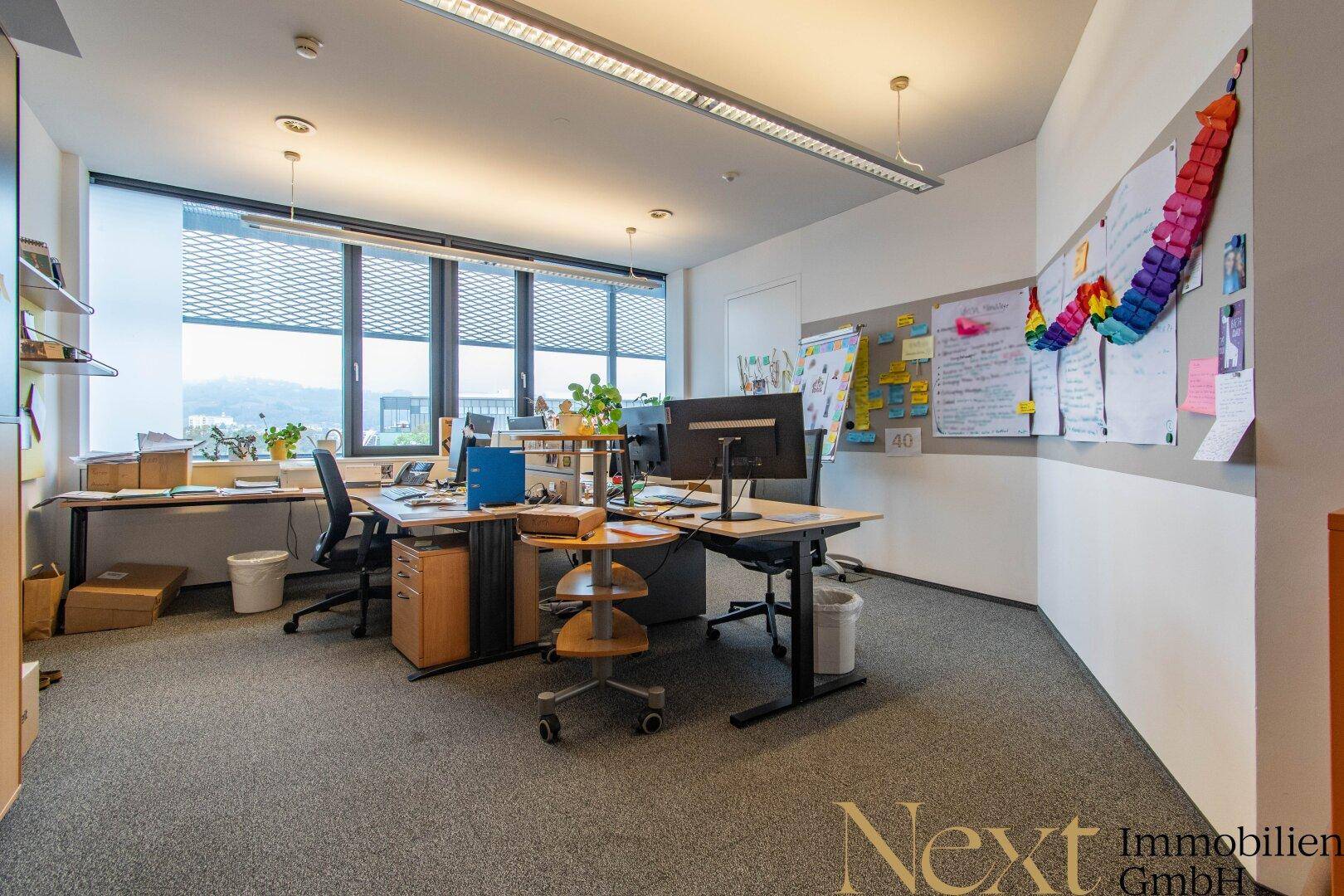 Geräumiger Bürobereich mit mehreren Arbeitsplätzen, Teppichboden und viel Tageslicht durch große Fenster.
