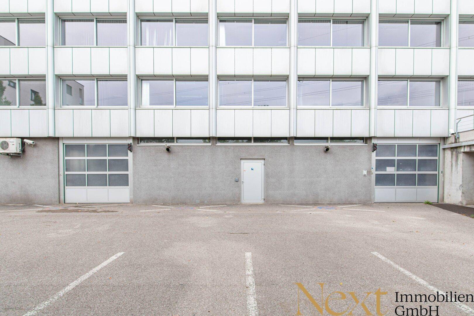 Rückseite des Gebäudes mit Garagentoren und einem Außenparkplatz für Fahrzeuge.