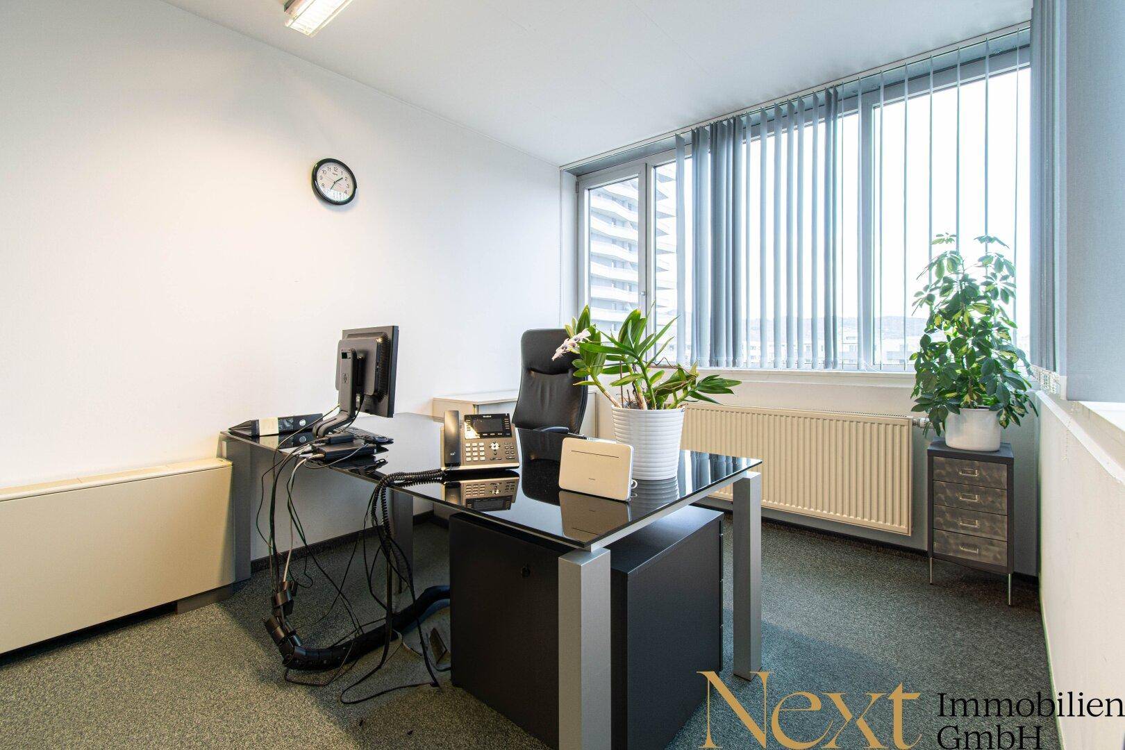 Helles Einzelbüro mit Schreibtisch, Bürostuhl und großem Fenster mit Jalousien, das viel Tageslicht hereinlässt.