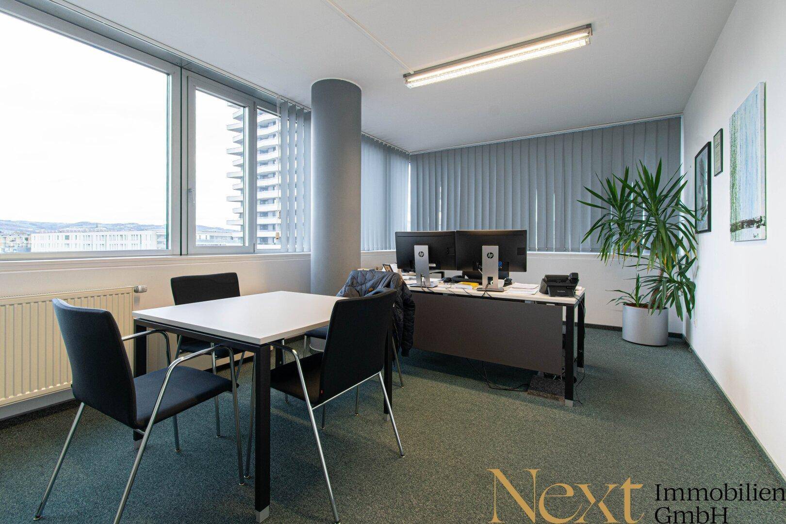 Helles und geräumiges Büro mit mehreren Arbeitsplätzen, großen Fenstern und Blick auf die Stadt, ausgestattet mit Teppichboden.