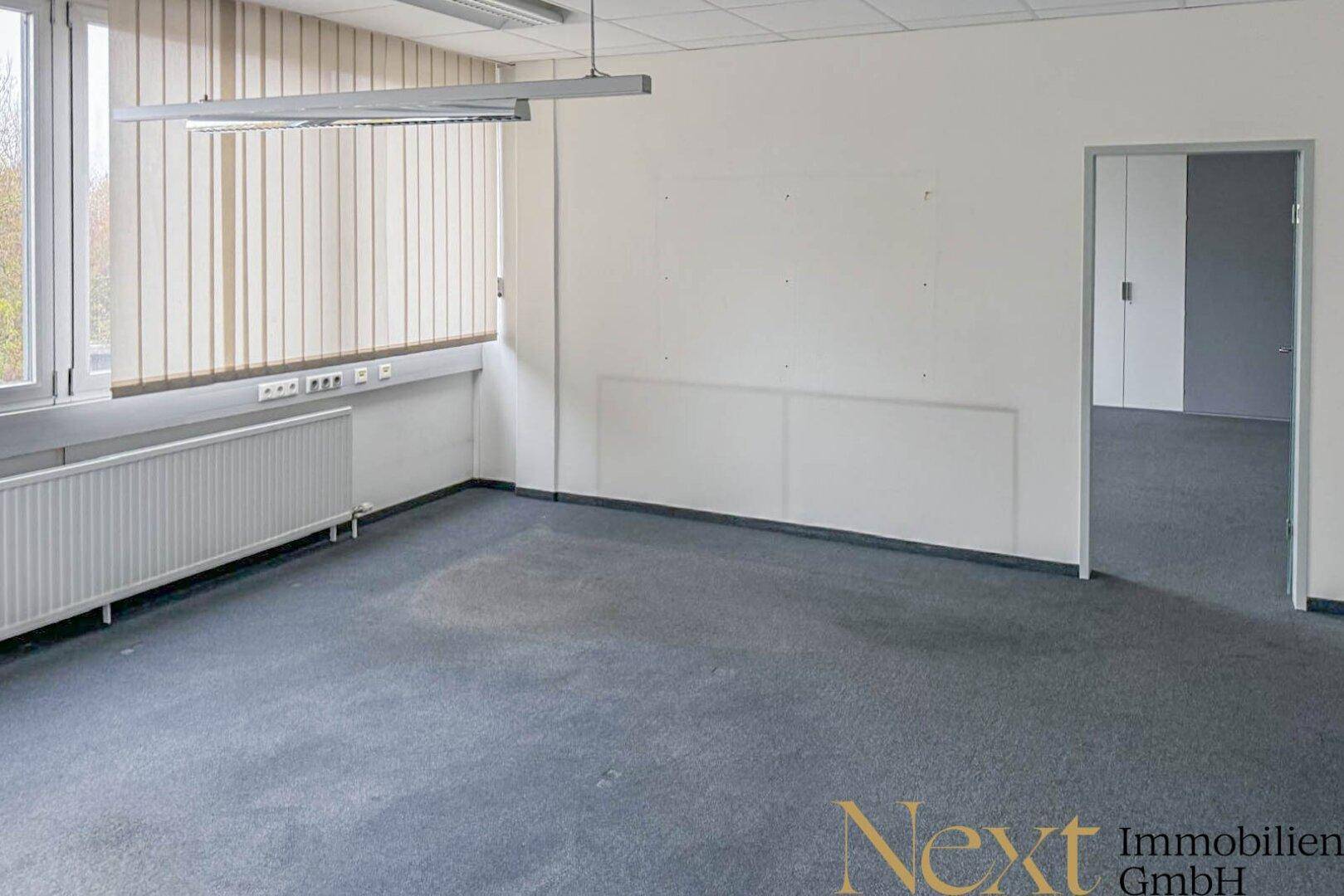 Leeres Büro mit Teppichboden, großen Fenstern und Zugang zu einem weiteren Raum.