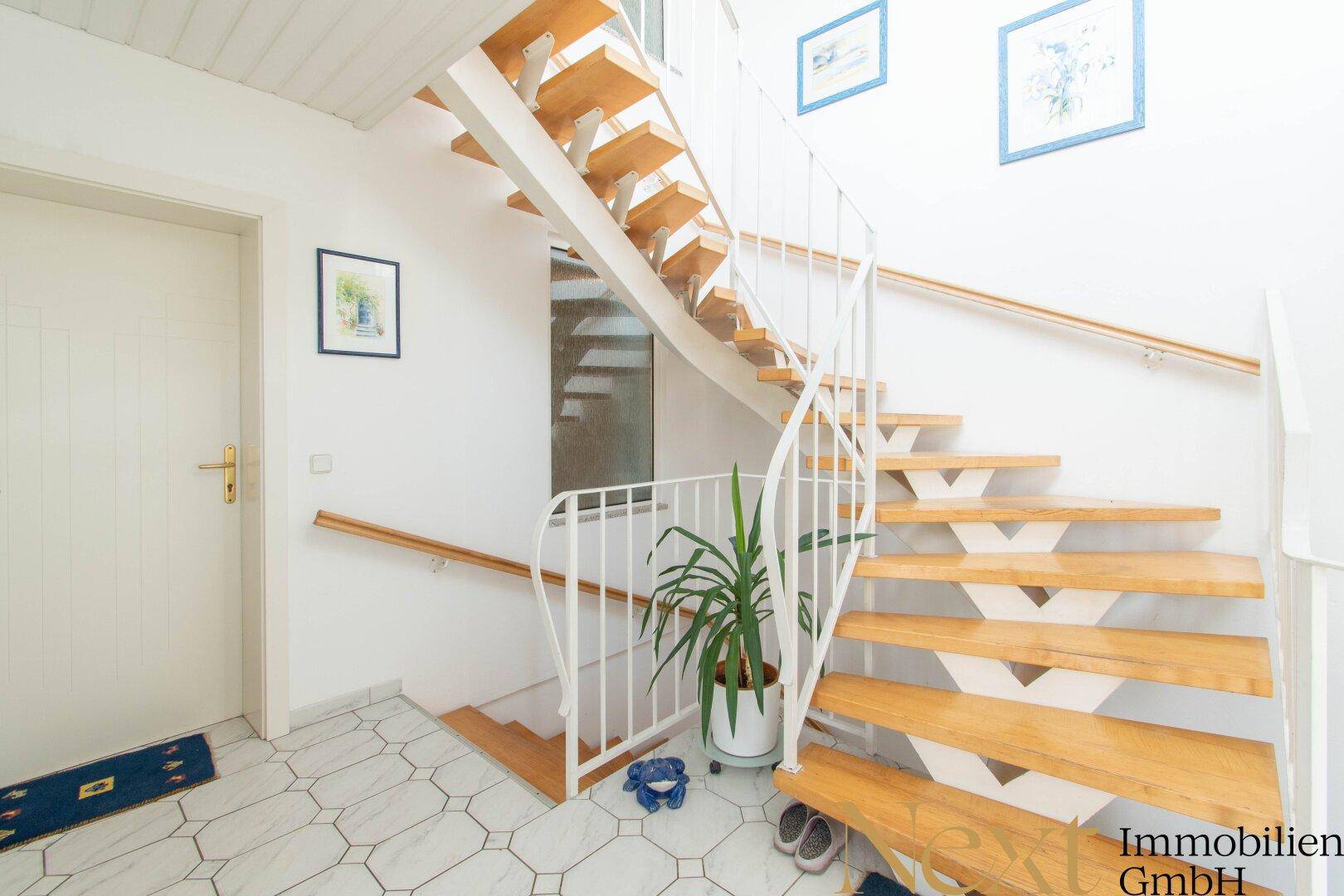 Heller Eingangsbereich mit eleganter Holztreppe, weißem Geländer und gemusterten Fliesen.