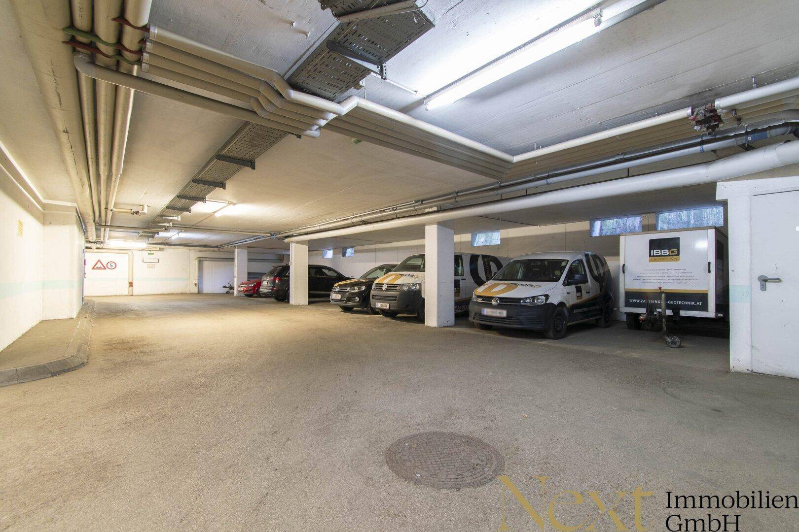 Geräumige Tiefgarage mit mehreren Stellplätzen, sichtbaren Leitungen an der Decke und Betonboden.