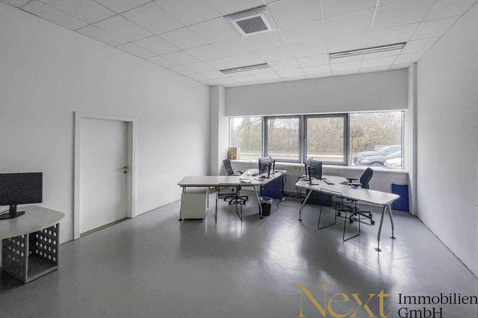 Helles Büro mit mehreren Schreibtischen, grauem Boden und großen Fenstern für viel Tageslicht.