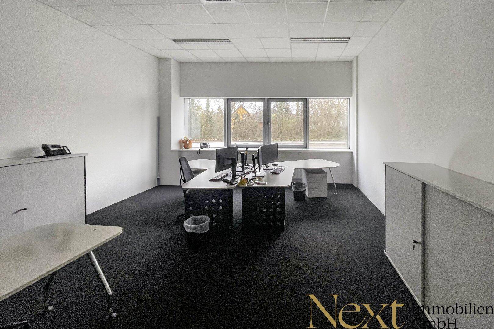 Modernes Büro mit mehreren Arbeitsplätzen, Teppichboden und großen Fenstern mit Blick ins Grüne.