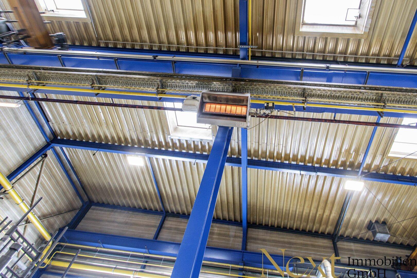 Blick zur Decke einer Industriehalle mit blauen Stahlträgern, Wellblechdach und einer Heizstrahleranlage.