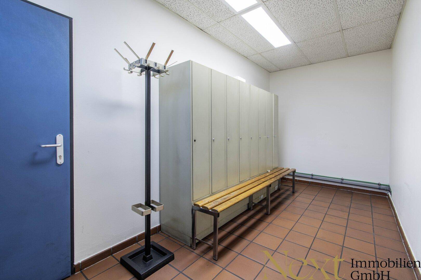 Umkleideraum mit Spinden, einer Holzbank und einem Garderobenständer auf Terrakotta-Bodenfliesen.