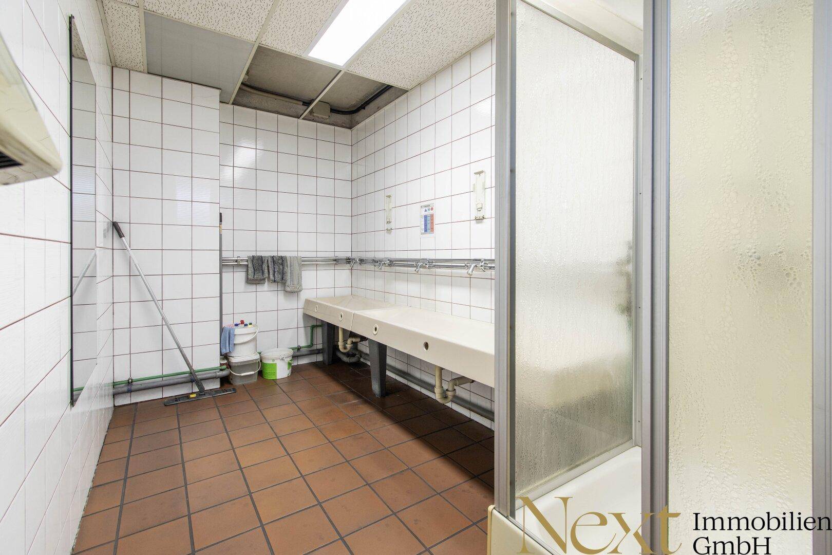 Waschraum mit mehreren Waschbecken, einer Duschkabine und weißen Wandfliesen.