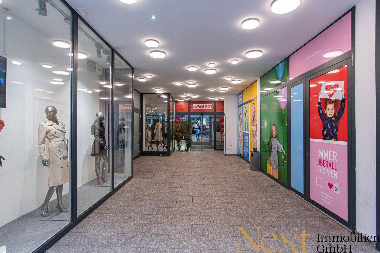 Helle und moderne Ladenpassage mit großen Schaufenstern, zahlreichen Deckenleuchten und robustem Steinboden.