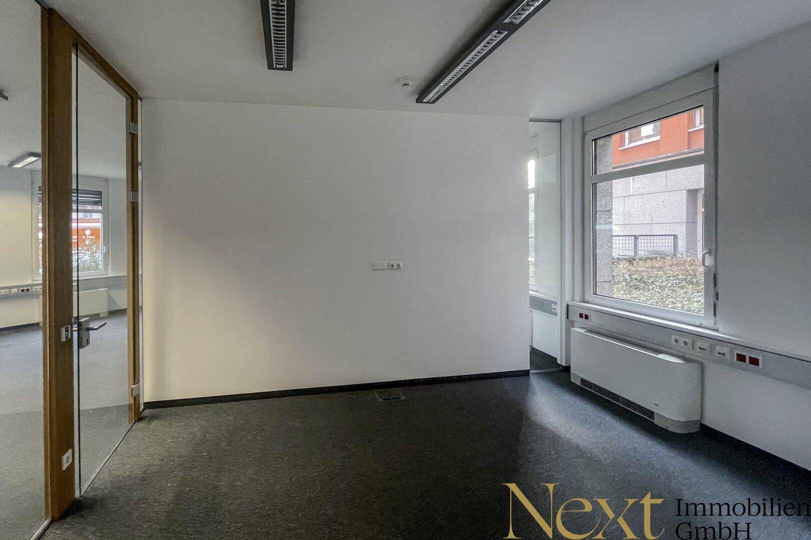 Leerer Bürobereich mit großen Fenstern, Teppichboden und einer Glasabtrennung, die den Raum strukturiert.