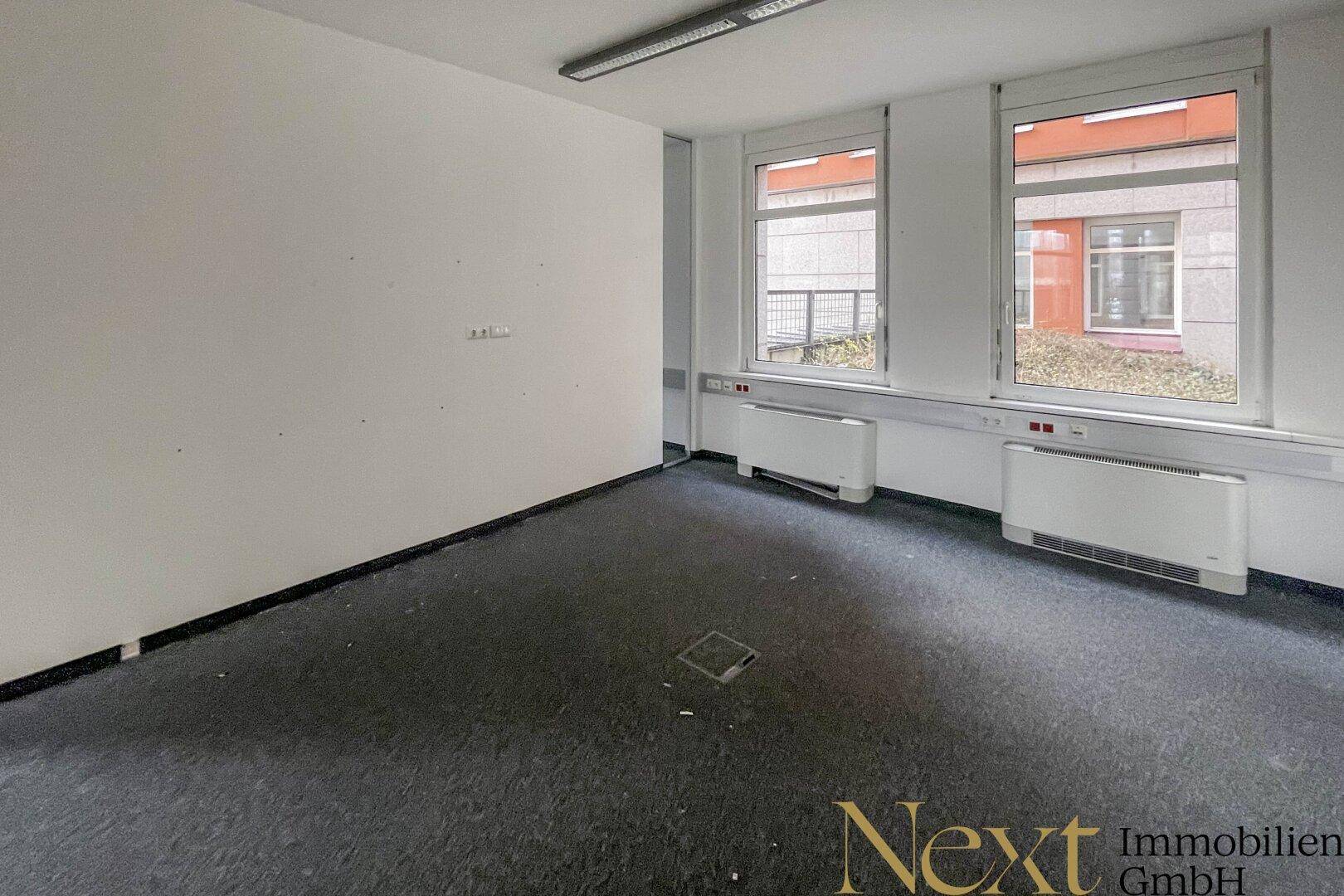 Heller Büroraum mit zwei großen Fenstern, Teppichboden und einer schlichten, funktionalen Ausstattung.