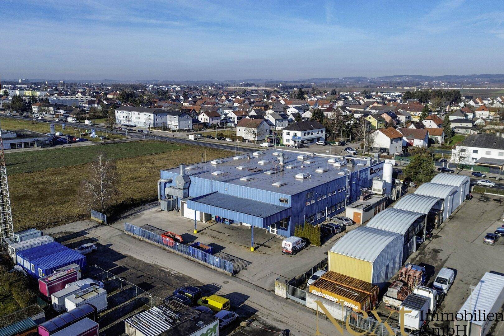 Luftaufnahme eines blauen Industriegebäudes mit Flachdach, umgeben von Lagerflächen und einem Wohngebiet im Hintergrund.