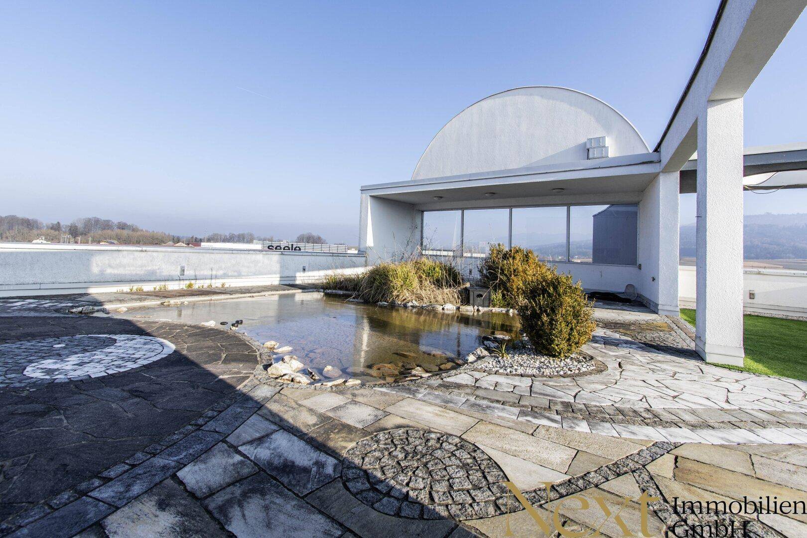 Große Dachterrasse mit dekorativen Steinplatten, einem Teich und Blick auf die Umgebung.
