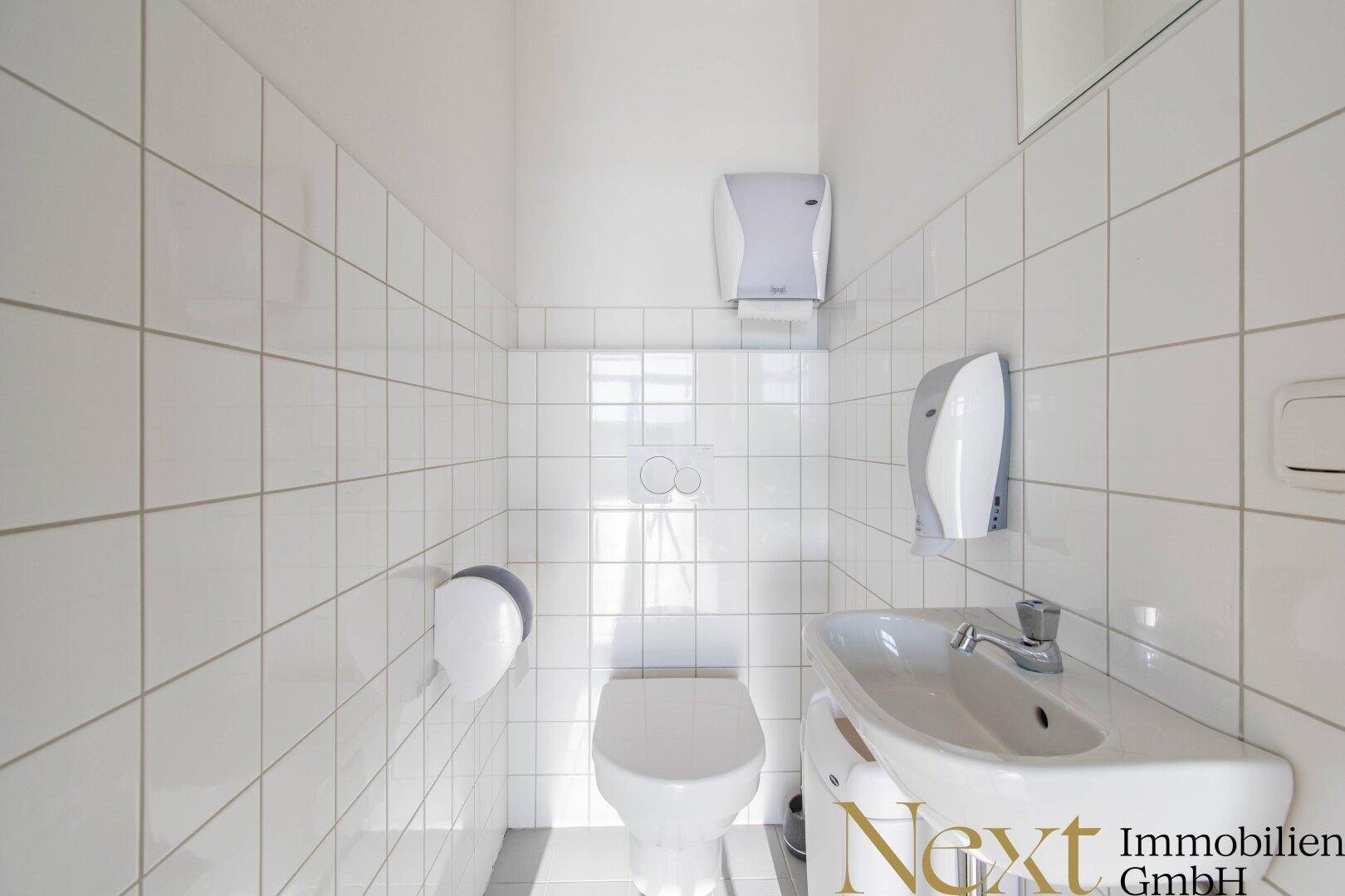 Einfache Gästetoilette mit weißen Fliesen, wandmontierter Toilette und kleinem Waschbecken.