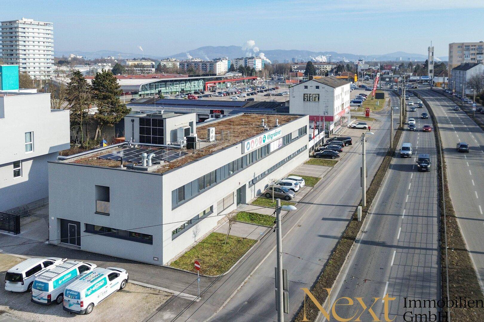 Luftaufnahme eines modernen Geschäftsgebäudes mit Photovoltaikanlage auf dem Flachdach und umliegenden Parkplätzen.