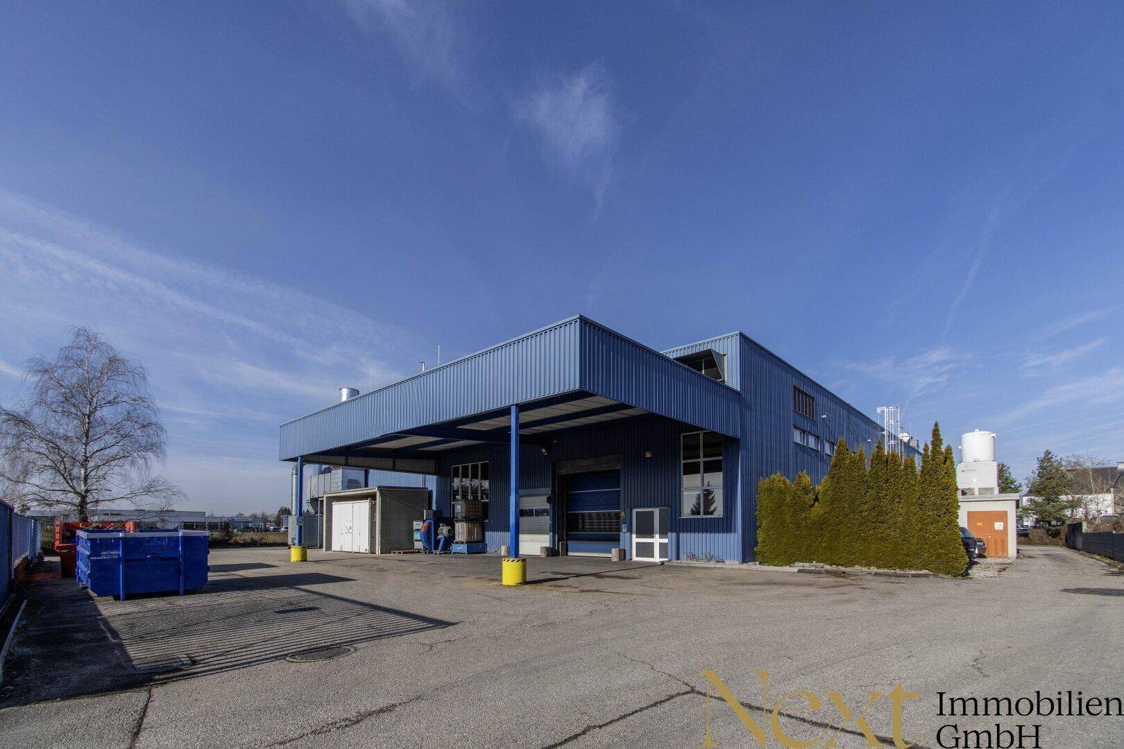 Ansicht des modernen Industriegebäudes mit blauer Fassade, Ladebereich und großem gepflasterten Vorplatz.