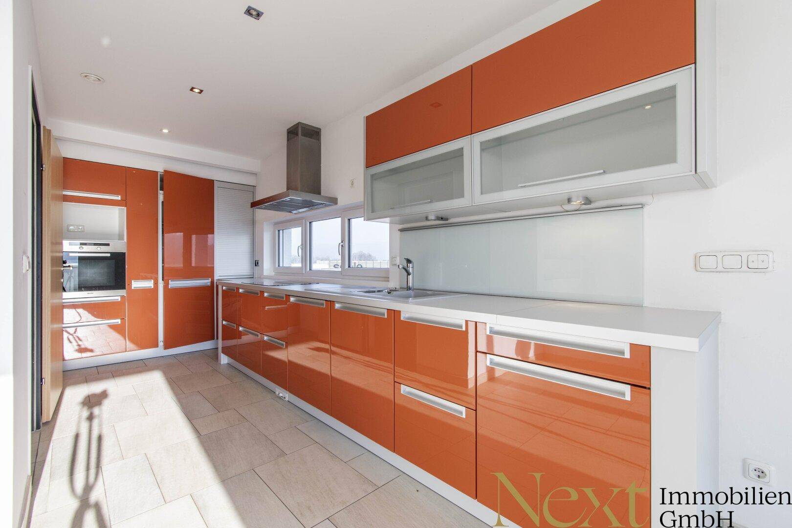 Moderne Küche mit glänzenden orangefarbenen Schränken, Edelstahlgeräten und Fenster.